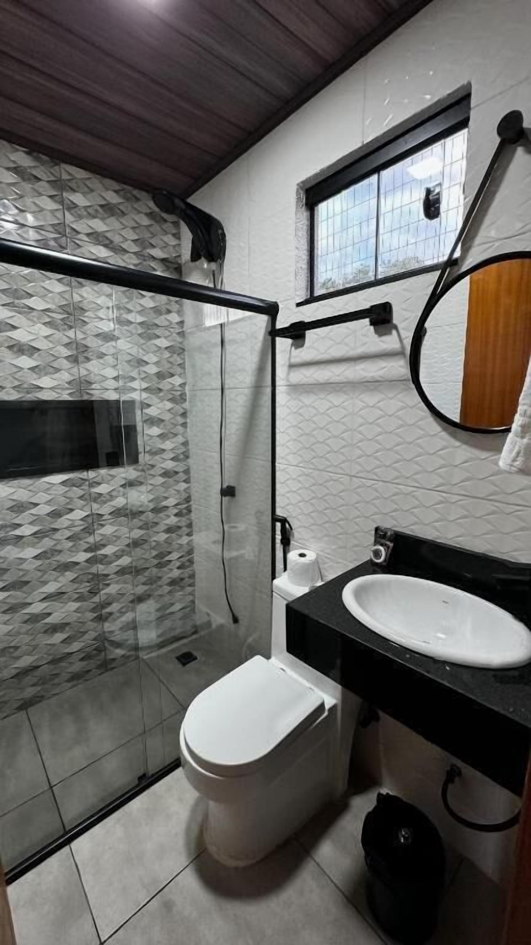 A bathroom with a glass shower enclosure, a white toilet, a sink with a black countertop, and a window with a view of the outside.