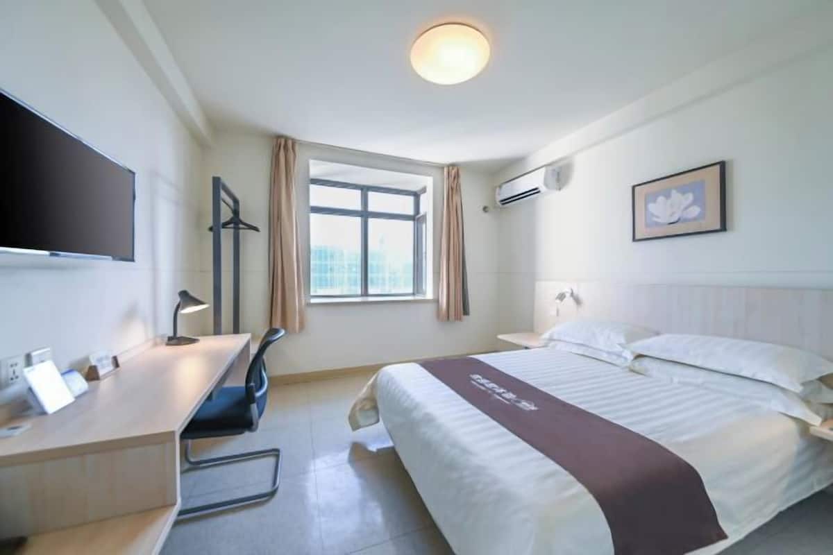 A hotel room with a bed, a desk, a chair, a TV mounted on the wall, a window with curtains, an air conditioning unit, and a framed picture on the wall.