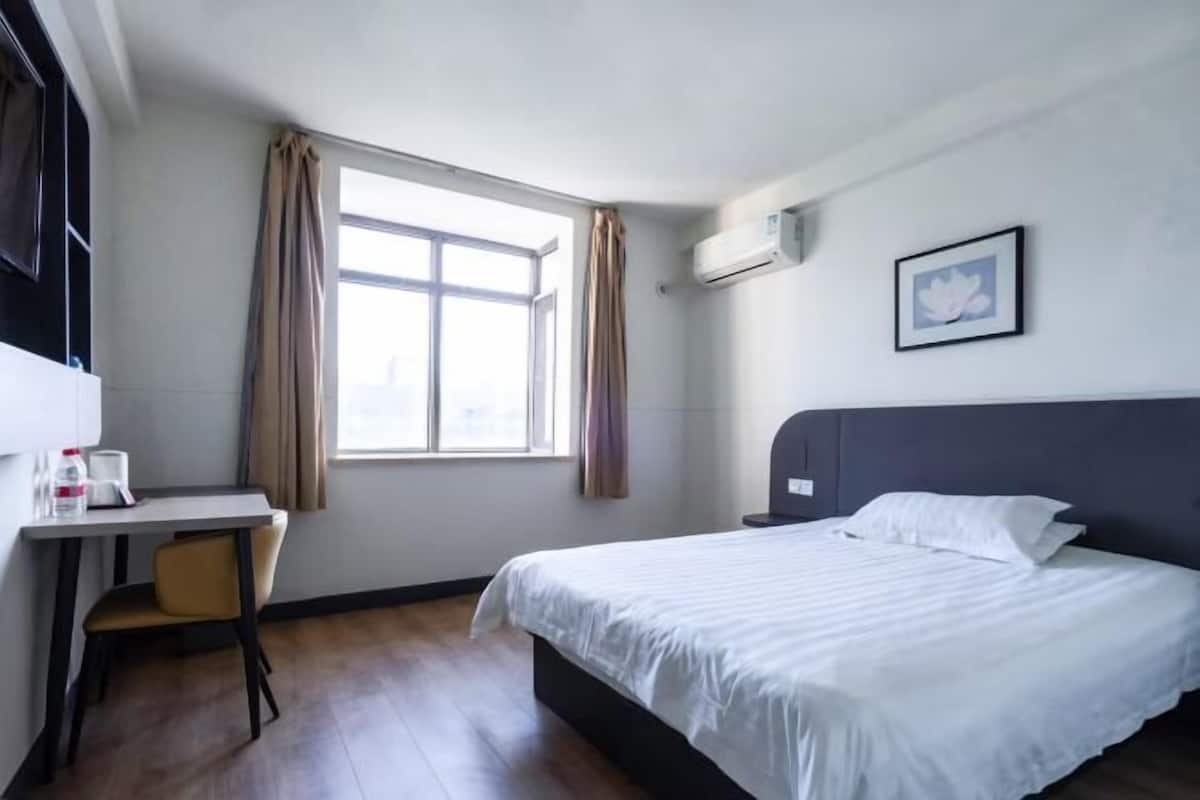 A hotel room with a bed, a desk, a chair, a window with curtains, an air conditioning unit, and a framed picture on the wall.