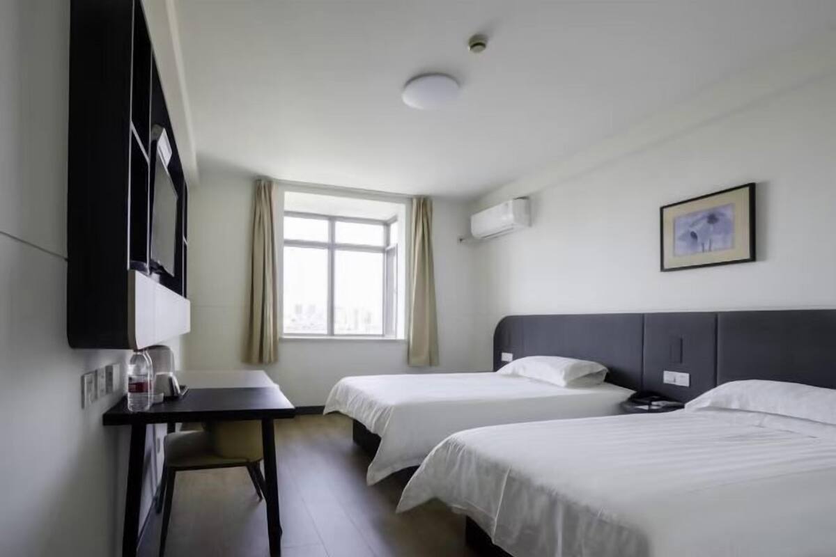 A hotel room with two single beds, a desk with a chair, a flat-screen TV, and a window with curtains.