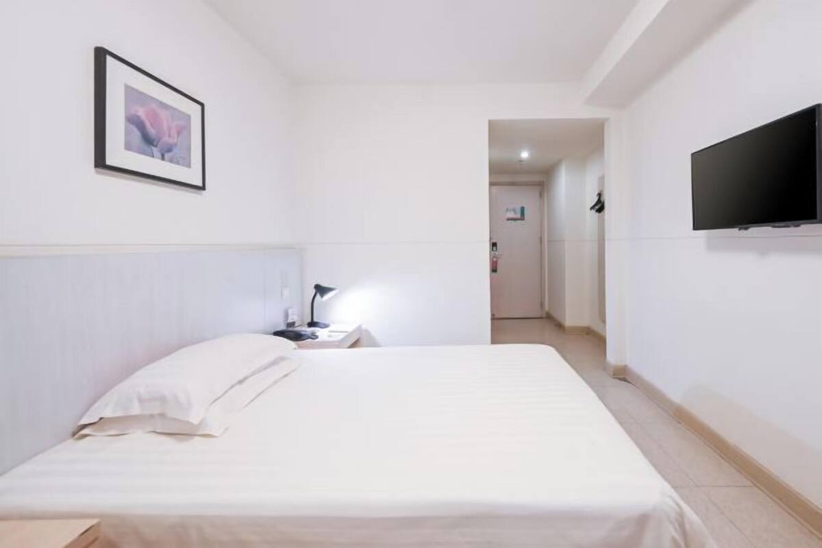 A hotel room with a bed, a television mounted on the wall, a framed picture on the wall, and a nightstand with a lamp.