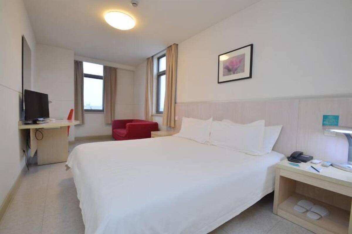 A hotel room with a large bed, a desk with a computer, a red chair, and a nightstand with a lamp and phone.