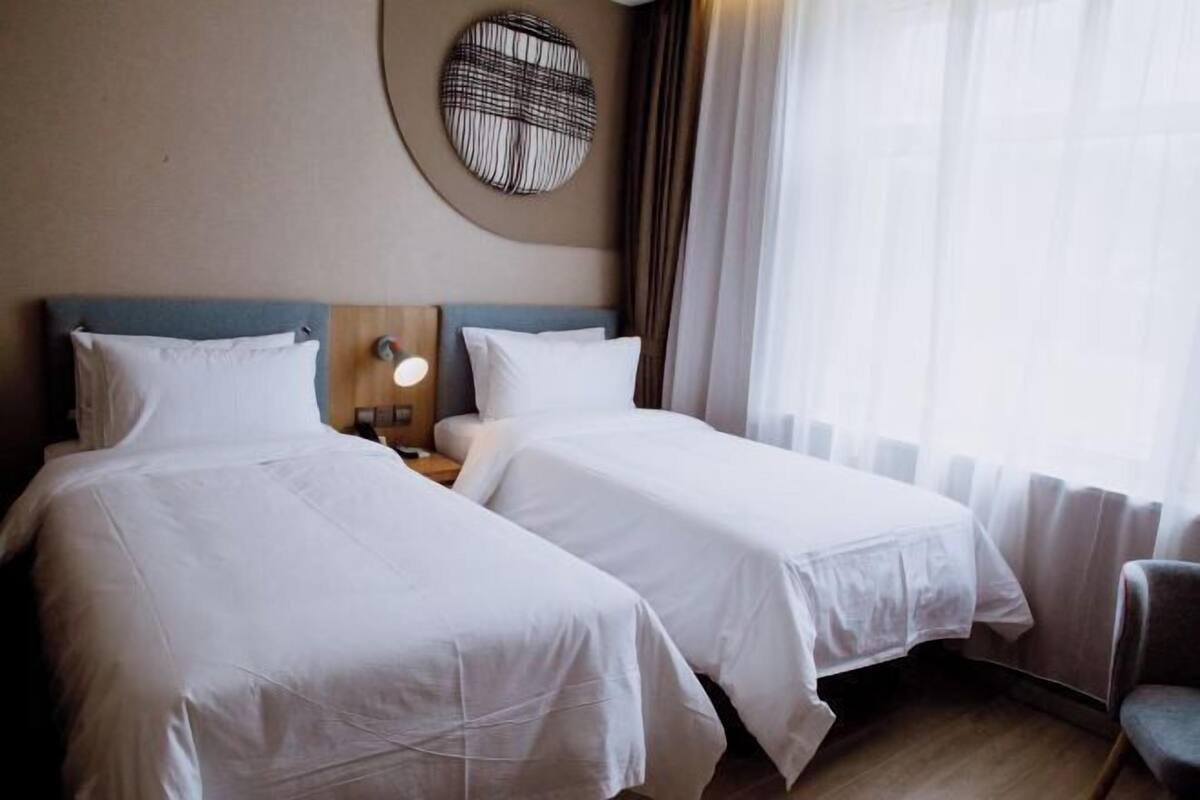 Habitación de hotel con dos camas, un adorno redondo en la pared, una mesita de noche con lámpara y vistas al exterior a través de cortinas translúcidas.