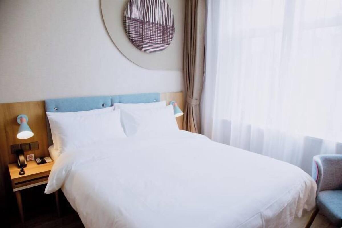 Una cama bien hecha con sábanas blancas, un adorno redondo en la pared, una lámpara de noche y una silla.