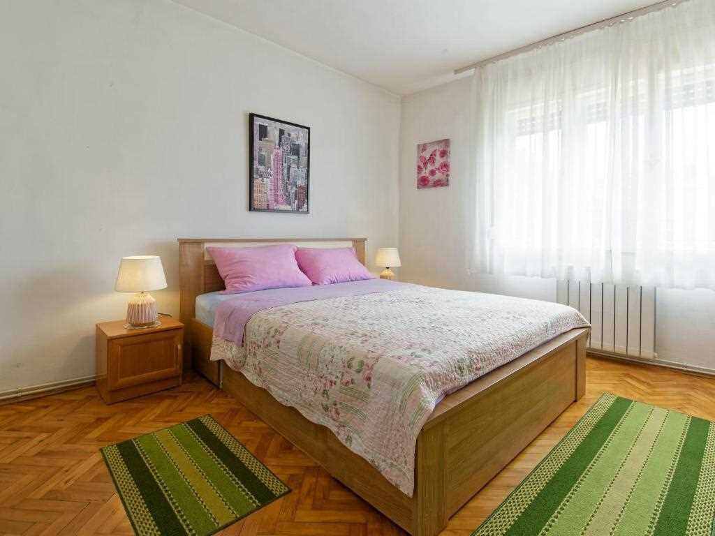 Ein Schlafzimmer mit einem hölzernen Bett, zwei Nachttischlampen, einem grün-weißen Streifen Teppich, einem Stadtbild an der Wand und einem Fenster mit transparenten Vorhängen.