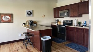 A kitchen with wooden flooring, a microwave, a stove, and a sink. There are stools and a trash can.