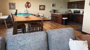 A living room with a wooden dining table, grey sofa, and a kitchen area with a microwave and cabinets.