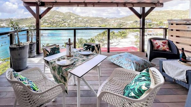 Une terrasse extérieure avec une table dressée pour deux personnes, entourée de chaises en osier et d’un banc rembourré.