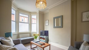 Een woonkamer met een bank, een bijzettafeltje met boeken en bloemen, een televisie en een raam met uitzicht op gebouwen.