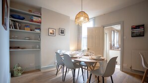 Een eetruimte met een ronde tafel gedekt voor zes personen, omringd door grijze stoelen, een hanglamp en een boekenkast met diverse spullen.