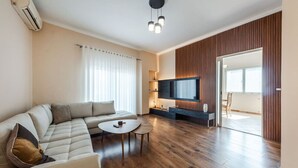 A modern living room with a sectional sofa, a round coffee table, a flat-screen TV mounted on a wooden panel wall, and a sliding glass door leading to a balcony. - The Old-Clock Apartment by PikHost (Shkodër)
