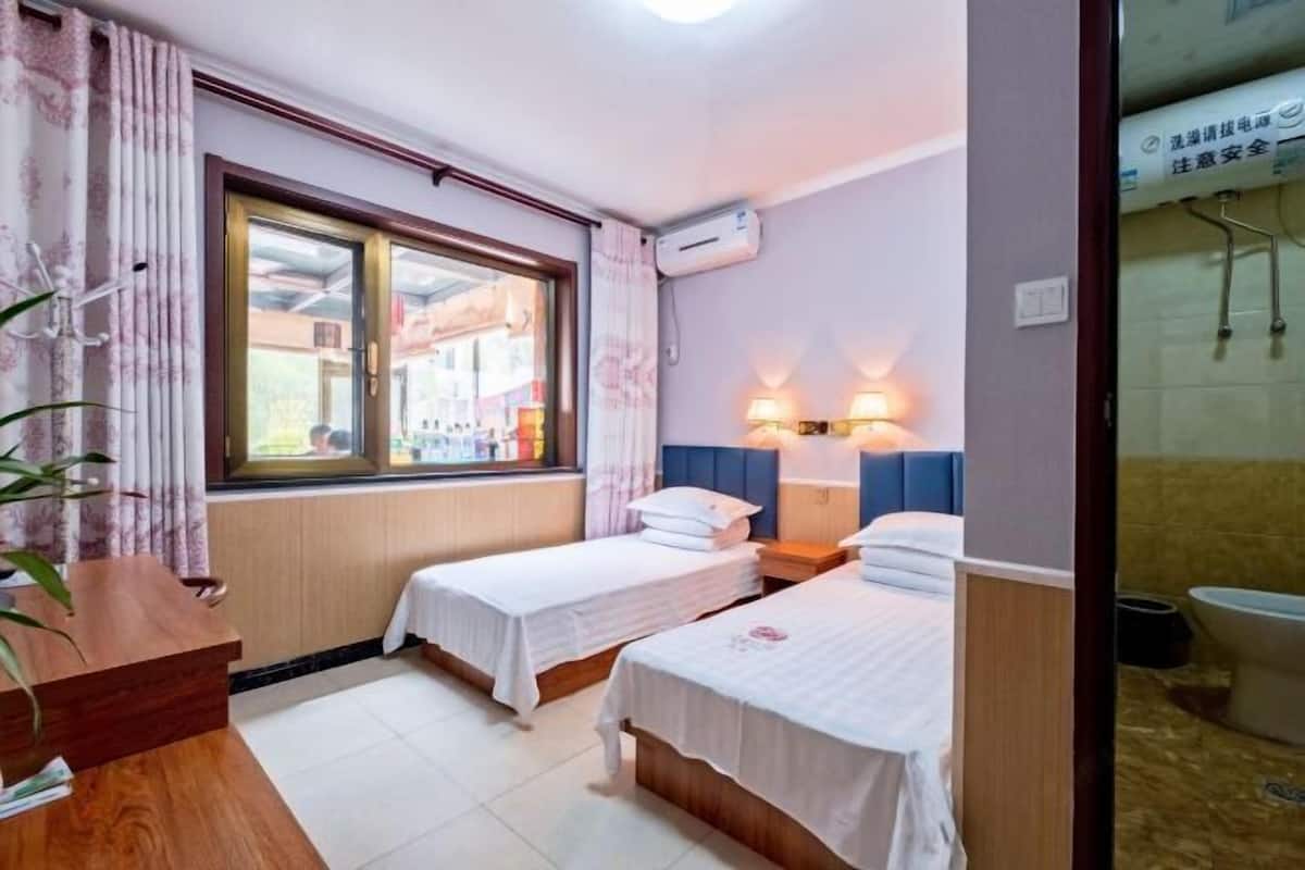 A hotel room with two single beds, a wooden desk, a window with curtains, an air conditioning unit, and a view of the street outside.