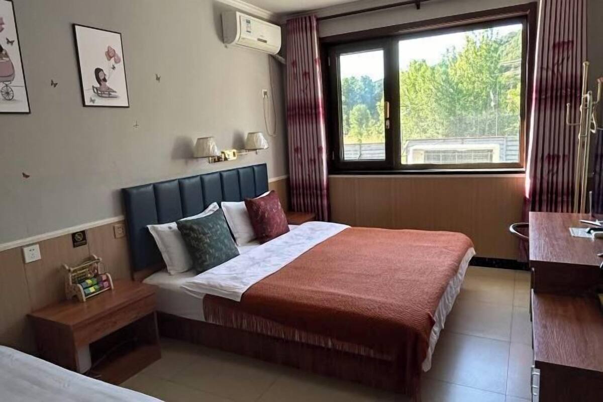 A hotel room with a bed, bedside table, lamp, and a window with a view of greenery.