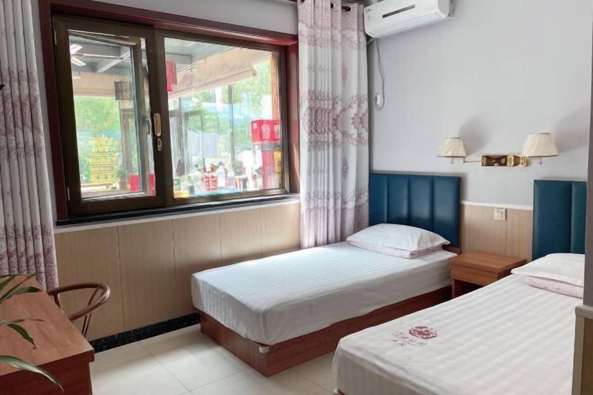 A hotel room with two beds, a window with curtains, a small table, and a wall-mounted air conditioner.