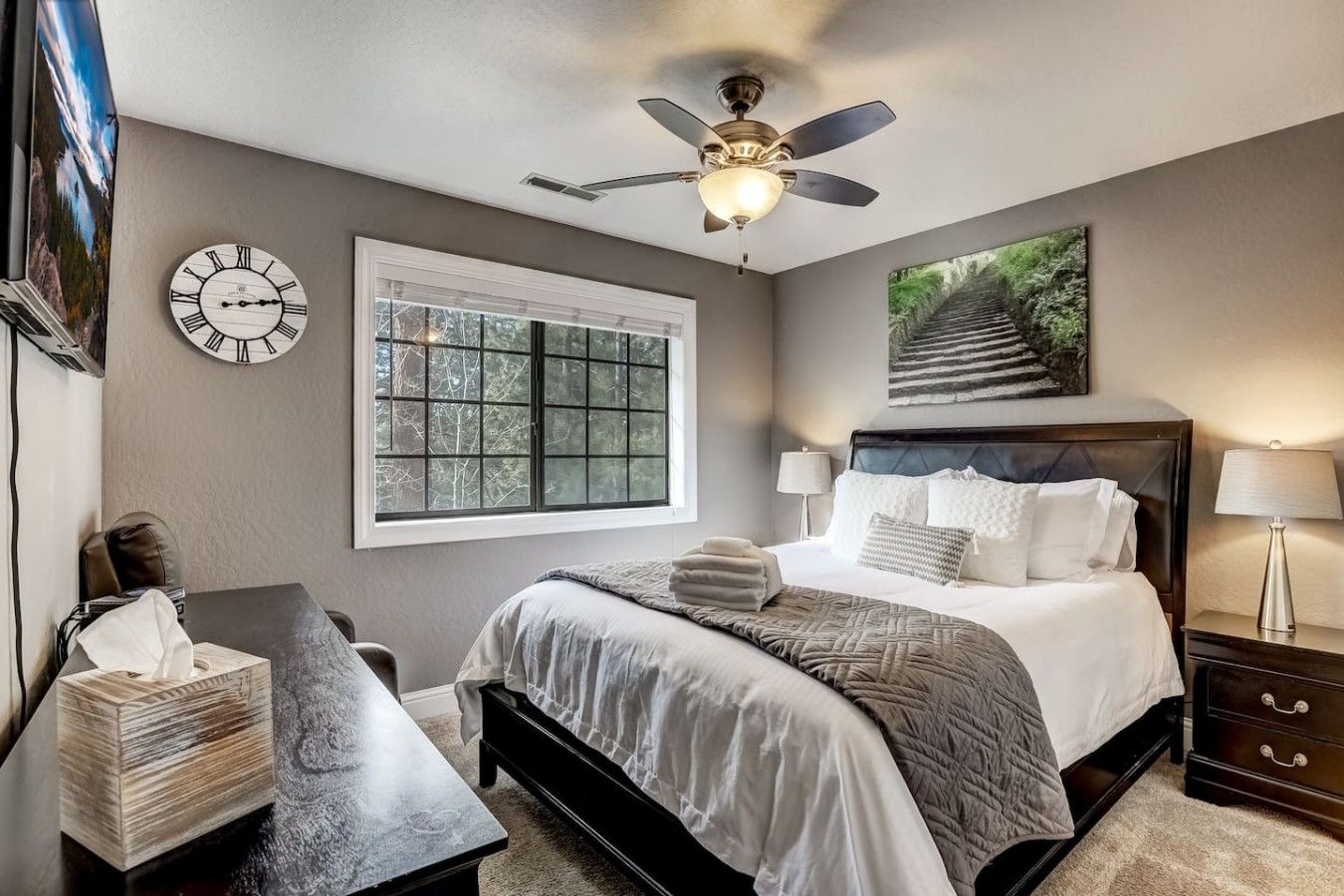 A bedroom with a bed, bedside tables, a window, a ceiling fan, and a clock on the wall.