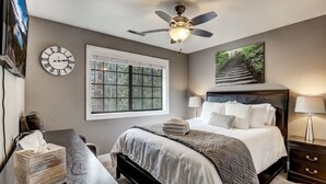 Een slaapkamer met een bed, nachtkastjes, een raam, een plafondventilator en een klok aan de muur.