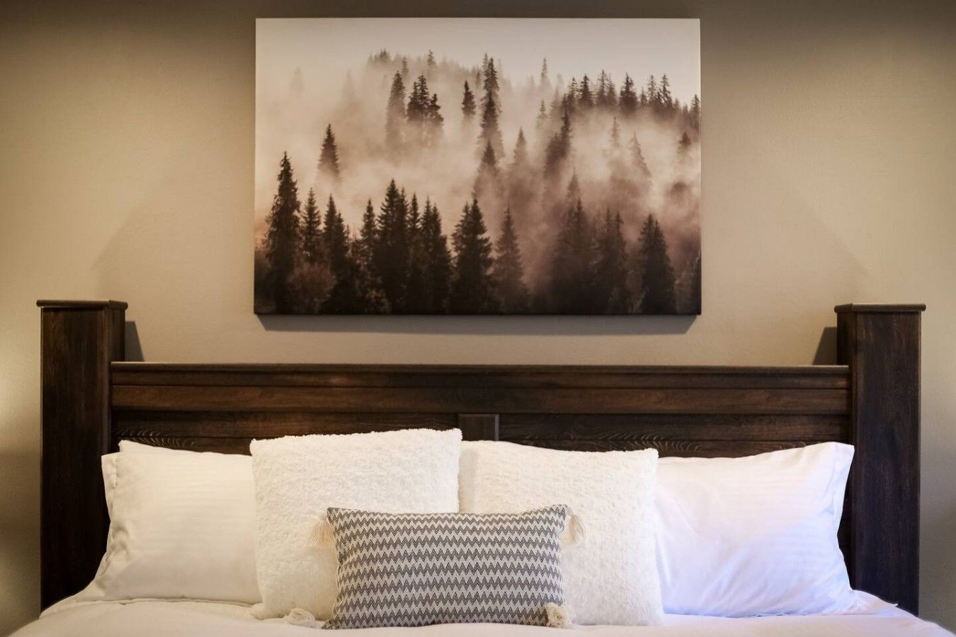 Una cama con ropa blanca y dos almohadas, una lisa y otra estampada, en una habitación con cabecera de madera oscura y un cuadro con motivos de bosque.