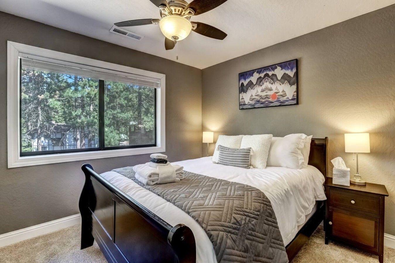 A bedroom with a bed, a nightstand, a lamp, a ceiling fan, a window with a view of trees, and a painting on the wall.
