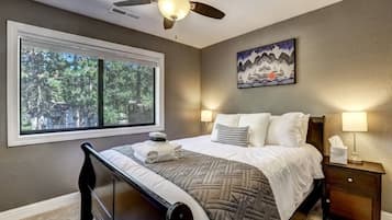A bedroom with a bed, a nightstand, a lamp, a ceiling fan, a window with a view of trees, and a painting on the wall.