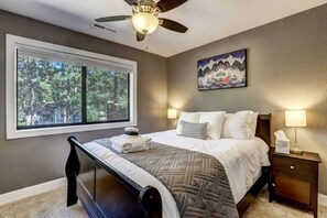Een slaapkamer met een bed, nachtkastje, lamp, plafondventilator, raam met uitzicht op bomen en een schilderij aan de muur.