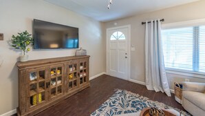 A living room with a flat-screen TV mounted on the wall, a wooden cabinet with glass doors, a door with a window, and a window with curtains. - Downtown Winter Garden bungalow with private putting green and fire pit (Winter Garden)