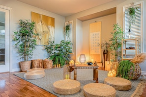 Una habitación con piso de madera, pufs tejidos, una mesa con velas y diversas plantas.