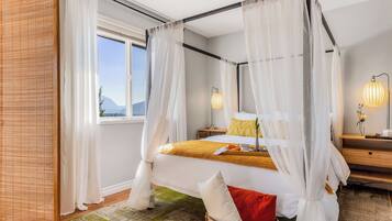 Una cama con dosel y cortinas traslúcidas, una mesita de noche con lámpara, y vistas a las montañas a través de una ventana.