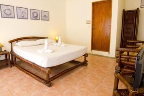 Una cama bien tendida con sábanas y toallas blancas, un cabecero de madera, una mesita de noche de madera con una lámpara, y un televisor sobre un mueble de madera.