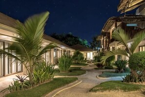 Une cour d’hôtel la nuit, avec des all ées illuminées, des palmiers et des chambres d’hôtes.