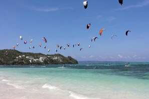 Una playa con agua turquesa cristalina, una orilla arenosa y una colina con edificios al fondo.