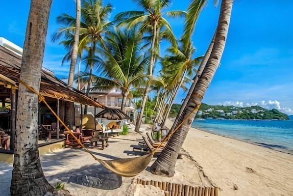 A beach with palm trees, a hammock, and a building with outdoor seating.