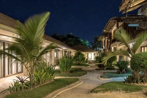 Un patio de hotel nocturno con senderos, palmeras y edificios con ventanas iluminadas.