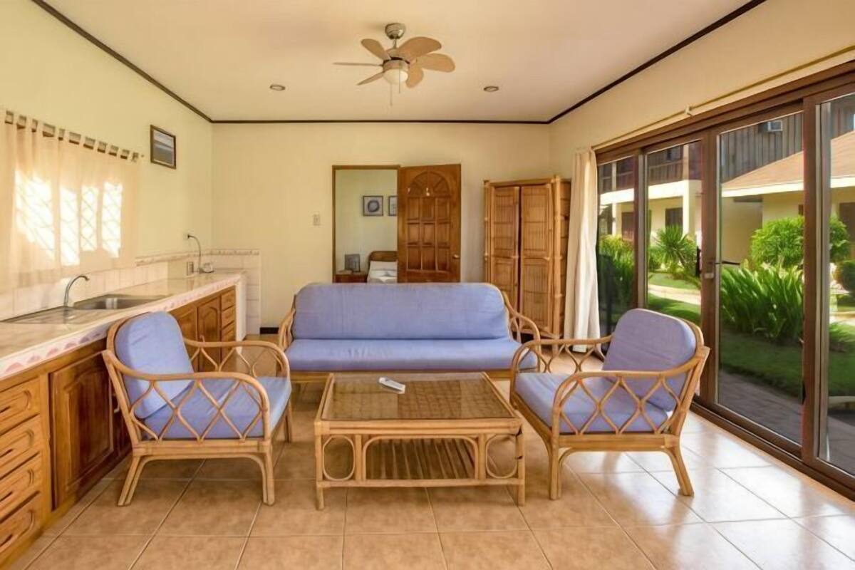 Une pièce dotée d’une kitchenette, d’un canapé bleu, de fauteuils en osier, d’un ventilateur de plafond et offrant une vue sur l’extérieur grâce à de grandes portes-fenêtres coulissantes.