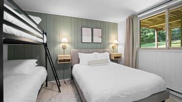 Un dormitorio con literas, una cama grande, dos mesitas de noche con lámparas, una ventana con vistas a la vegetación y dos cuadros en la pared.