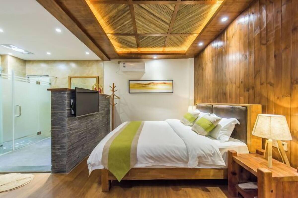 Un dormitorio moderno con techo de madera, una cama con ropa de cama blanca y detalles verdes, una mesita de noche con una lámpara y un televisor montado en una pared de piedra.