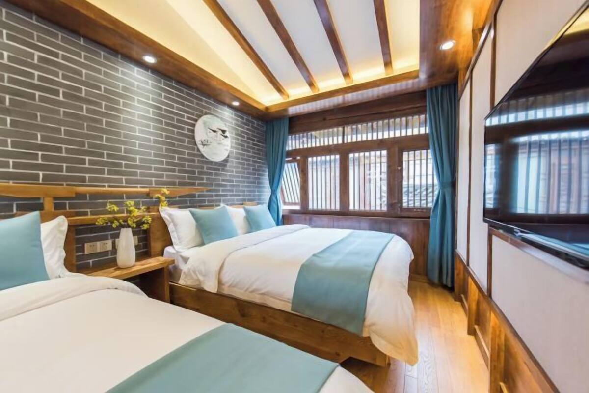 Habitación de hotel con dos camas, una pared de ladrillo, vigas de madera, un reloj y una ventana con cortinas.