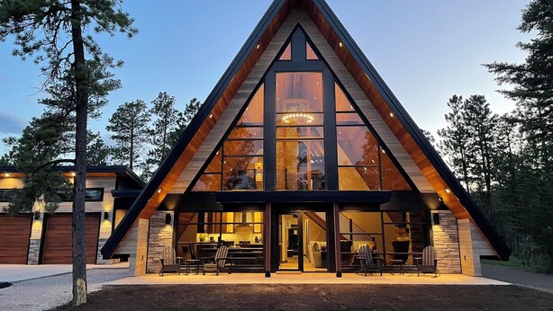 A modern A-frame lodge with large windows, a stone exterior, and a well-lit entrance area with outdoor seating.