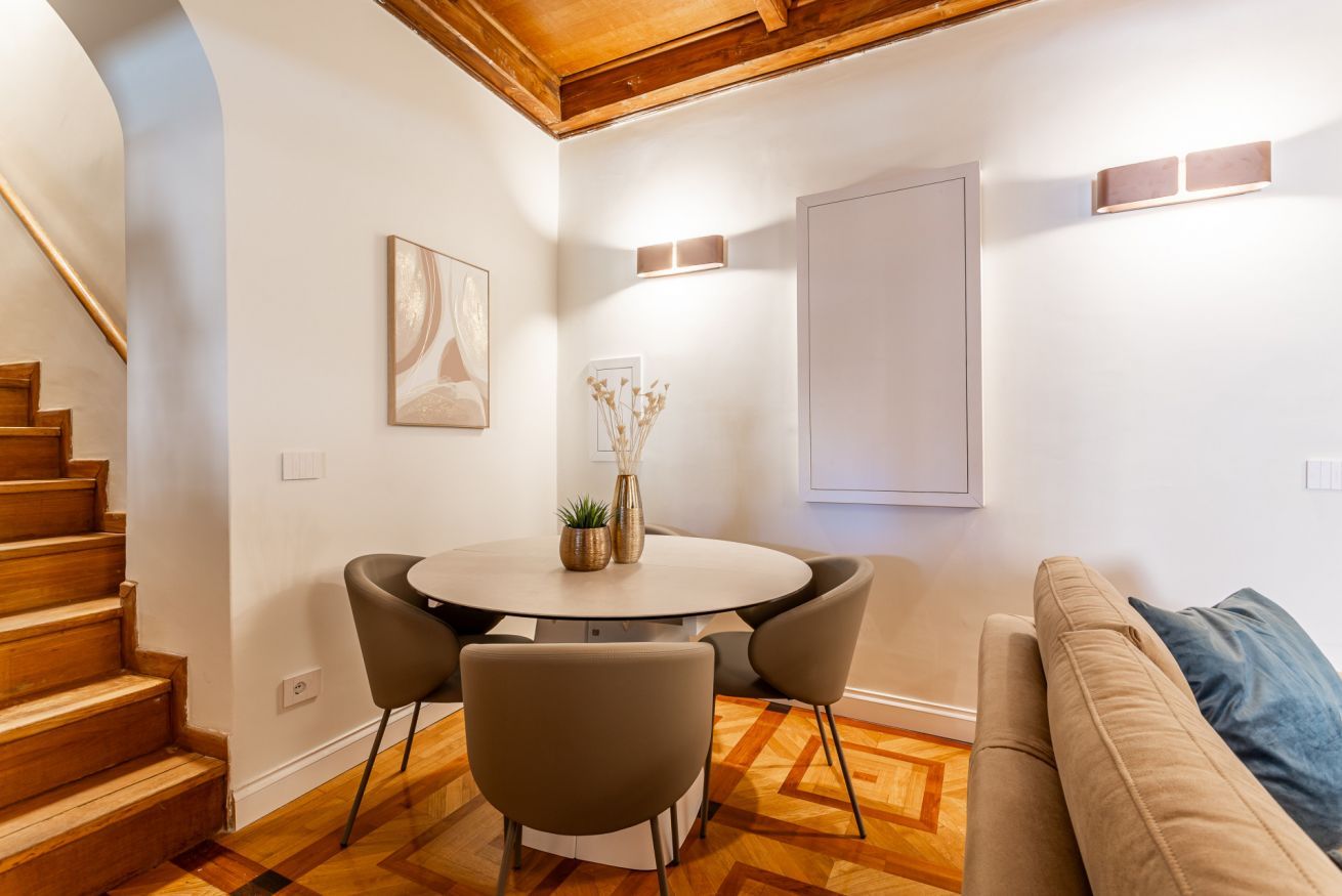 Een moderne woonkamer met een houten trap, een ronde eettafel met stoelen, een bank en een wandlamp.