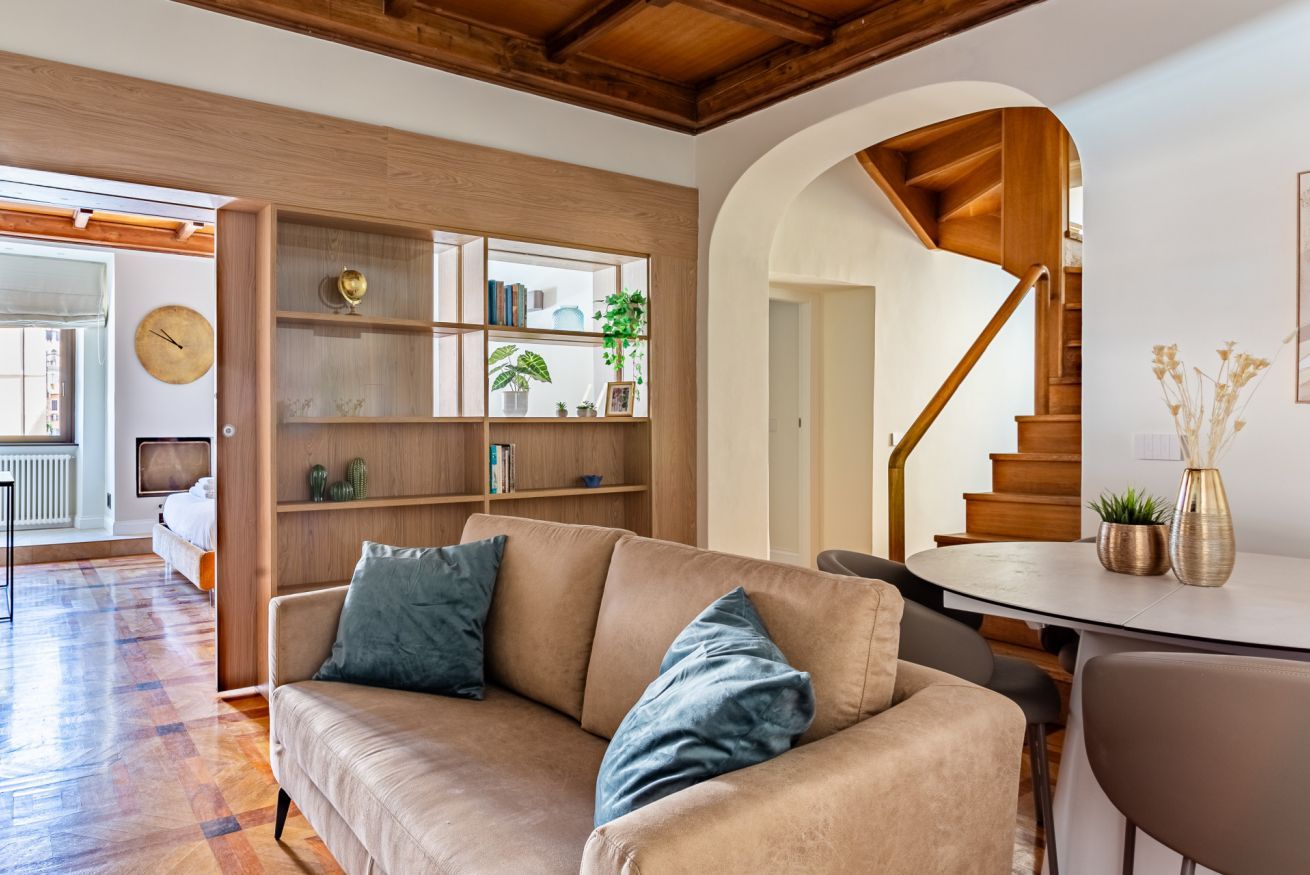 A living room with a sofa, a wooden bookshelf, a dining table, and a staircase.