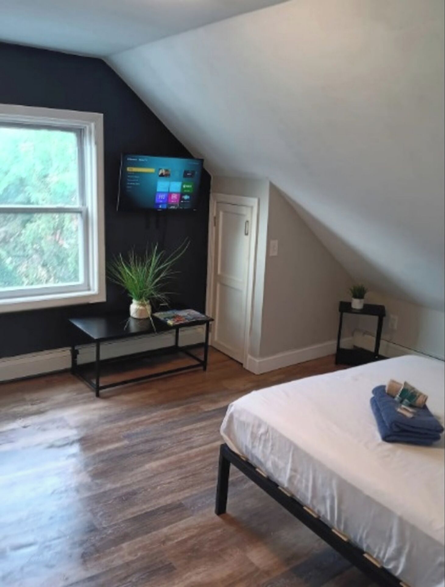 Un dormitorio con una cama, un televisor montado en la pared, una ventana y una mesita con una planta.