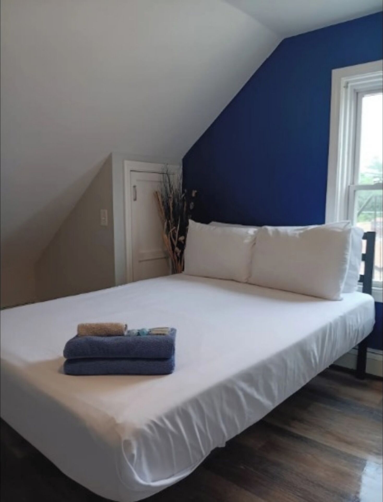 Une chambre avec un grand lit, du linge de lit blanc, un coussin bleu en guise d’élément décoratif, et une fenêtre donnant sur un beau paysage.