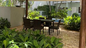 Een overdekt terras met een tafel en stoelen, omringd door weelderige begroeiing en een betegelde vloer.