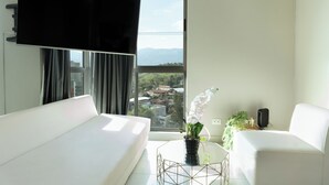 Une chambre d’hôtel moderne dotée d’une grande fenêtre, d’un téléviseur à écran plat, d’un canapé blanc, d’une petite table avec une plante et d’un fauteuil blanc.