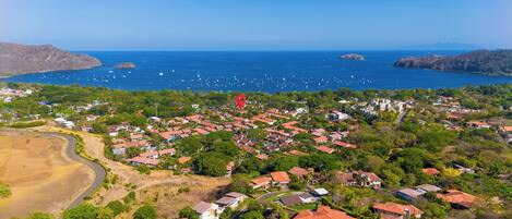 An aerial view of a coastal residential area with houses, a winding road, and a clear view of the ocean with boats.