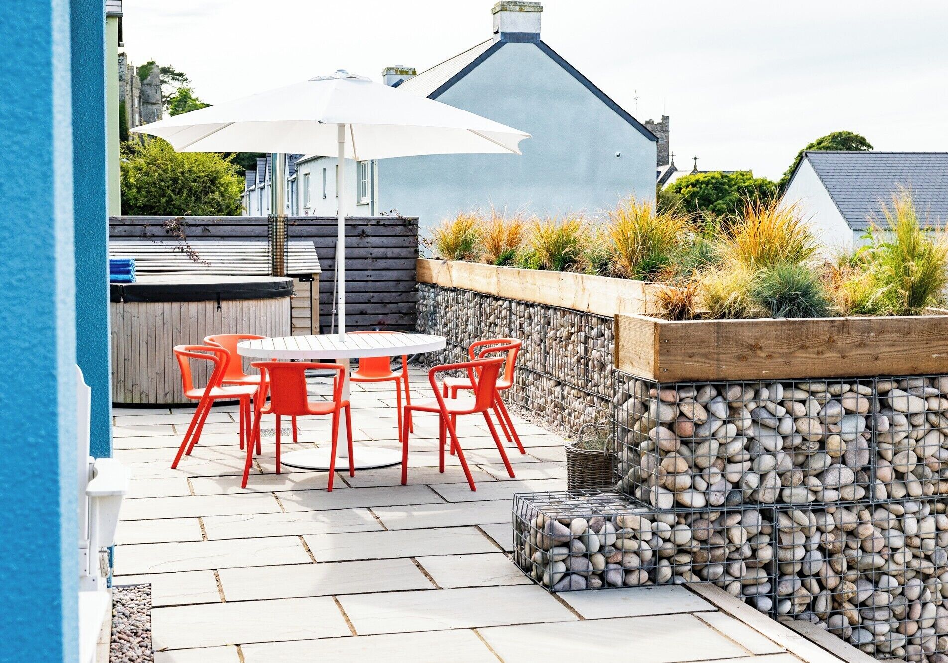 Eine Terrasse im Freien mit einem runden Tisch und roten Stühlen, einem weißen Sonnenschirm und einer Steinmauer mit Pflanzen.