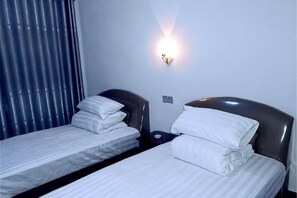 Deux lits simples avec des têtes de lit, une lampe fixée au mur et une fenêtre avec des stores.