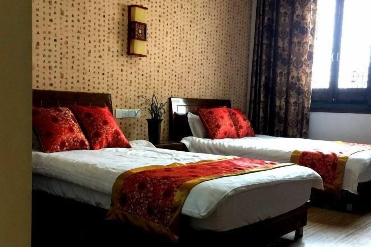 A hotel room with two beds, each with red pillows and white linens. There is a small table with a plant between the beds, and a window with curtains.