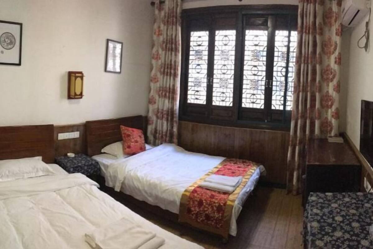 A room with two beds, a wooden headboard, a window with curtains, a wall-mounted clock, and a small table.