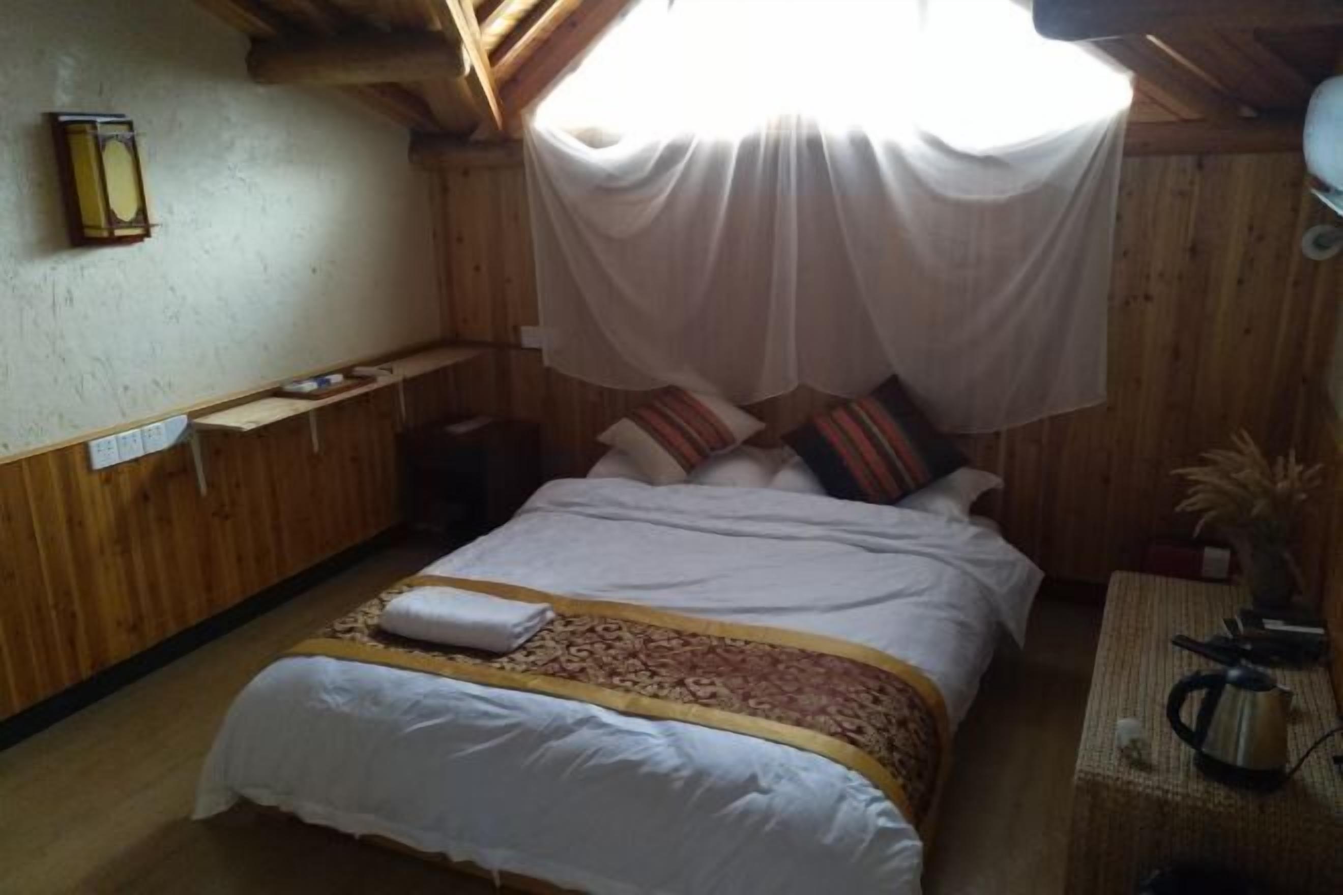 A wooden room with a bed, a nightstand, a lamp, and a window with a white curtain.