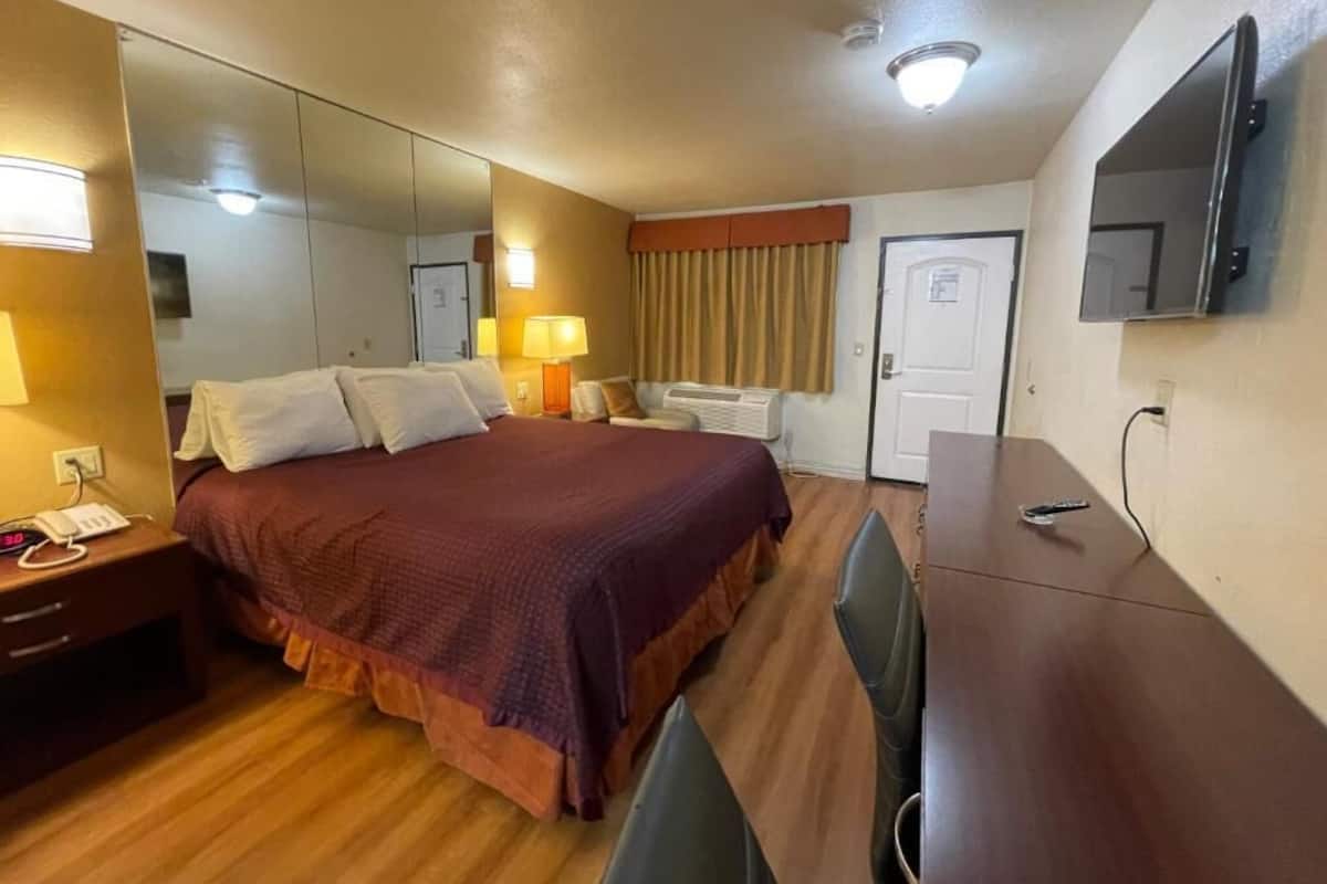 A hotel room with a bed, bedside table, lamp, mirror, TV, and a desk with chairs.